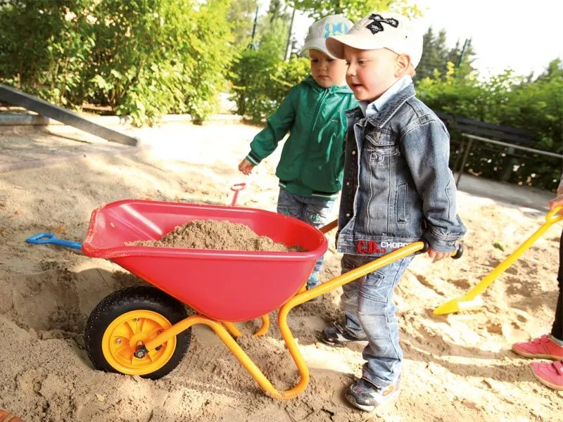 Kinderschubkarre gefüllt mit Sand und fröhlich spielenden Kindern im Sandkasten